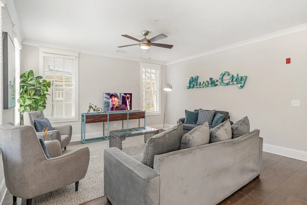The image captures a spacious and inviting living room with a plush sofa adorned with decorative pillows, a television displaying a vibrant live music performance, a bold 'Music City' wall sign, and lush green plants adding a touch of life. The scene exudes warmth and is perfect for relaxing, socializing with friends or family, or immersing in Nashville's musical spirit from the comfort of your stay.