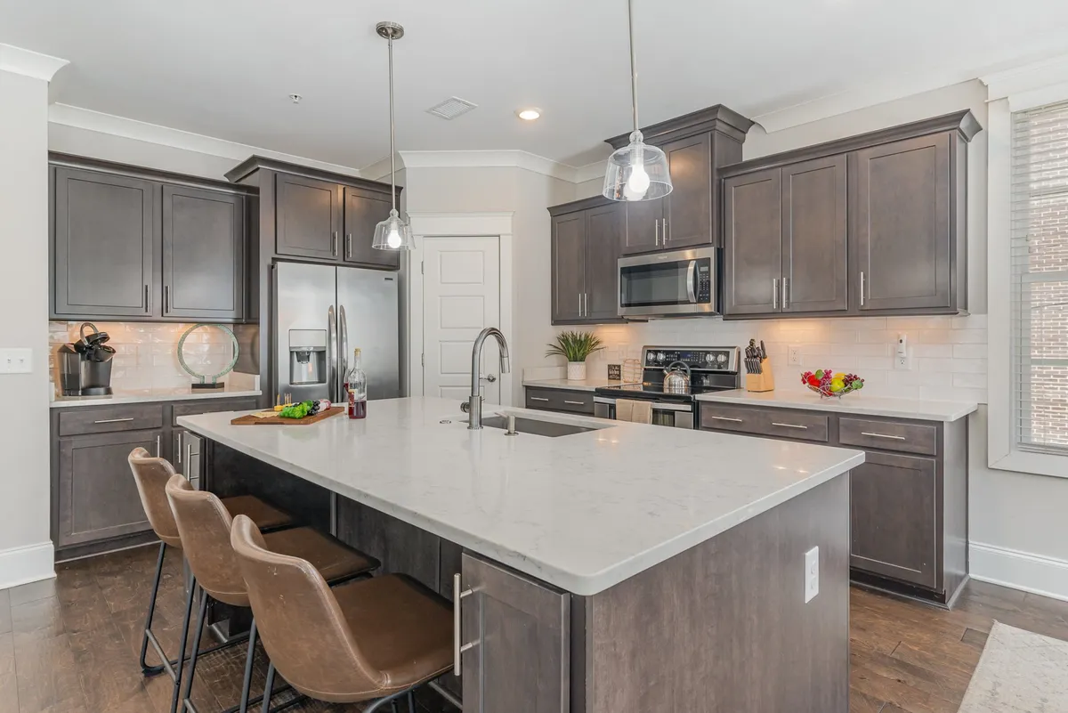 A bright, contemporary kitchen scene featuring a prominent kitchen island, bar stools, modern appliances, and elegant cabinetry. The space invites socializing, cooking, and casual dining, making it the heart of the home for guests.